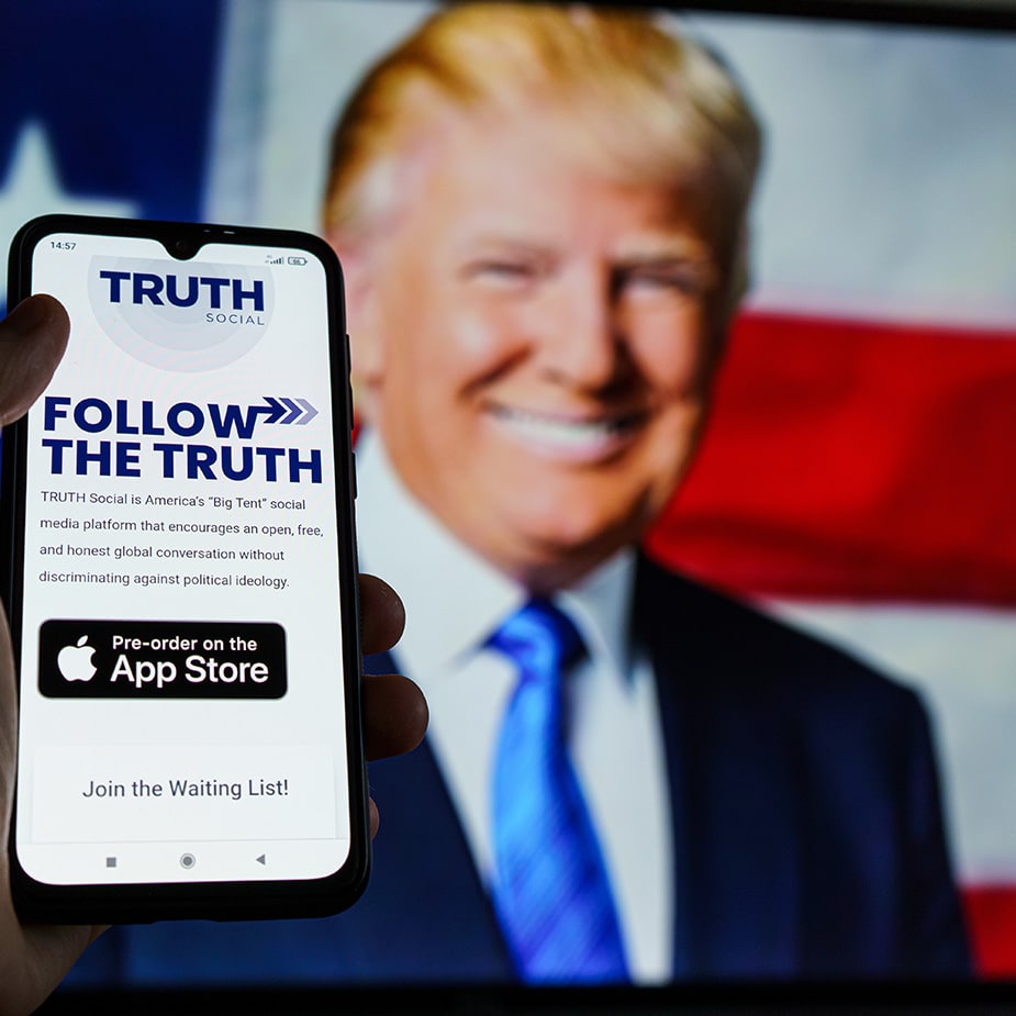 An image of a hand holding a mobile phone with the TRUTH Social app that symbolizes our TRUTH Social Ads services. In the background, there's an image of a smiling and radiant President Donald J Trump. TRUTH Social can be a great tool for Republican and Conservative political advertising campaigns.