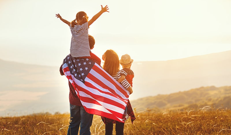 An image of a nuclear American family (father, mother, and two children) enjoying the sunset, with the dad holding an American flag.